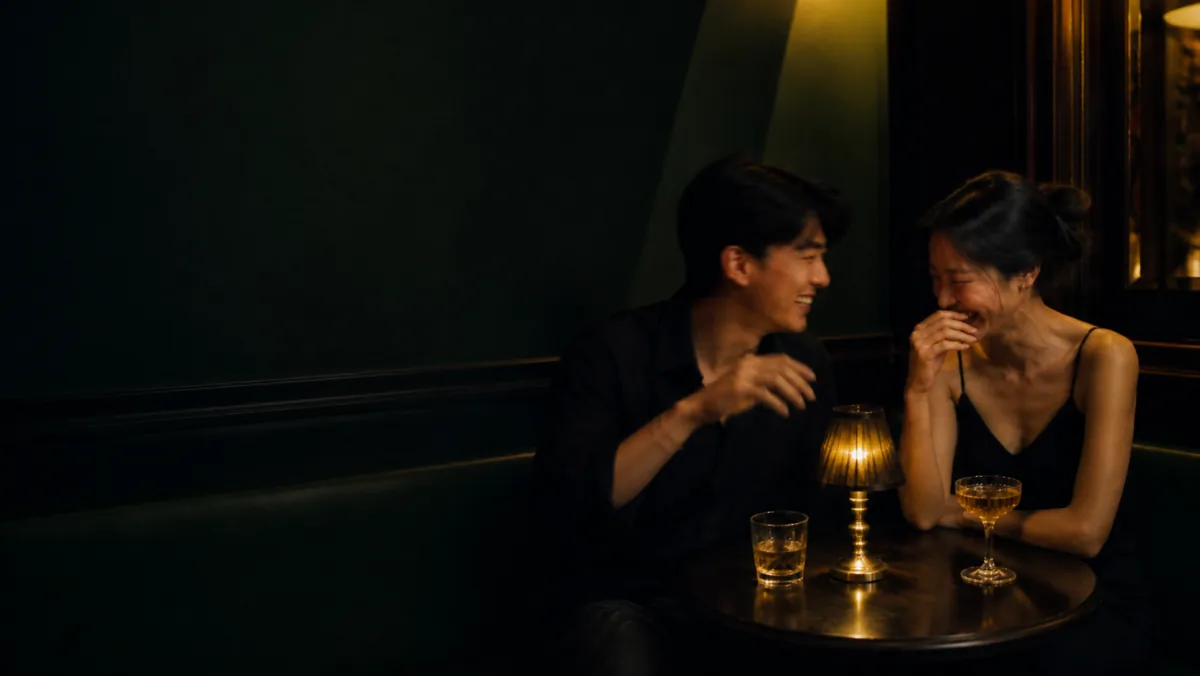 An Asian pair laughing over a coupe of wine in a dark cocktail bar.