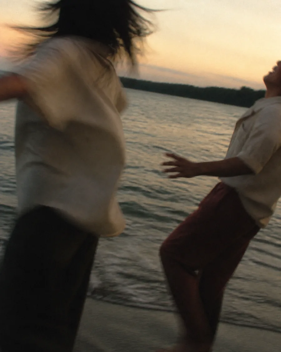 An Asian pair running and laughing together at the water’s edge near sunset.
